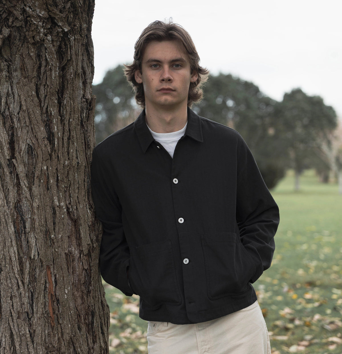 Model wearing the Anderson Chore Jacket in black over a white tee and cream trousers, leaning against a tree in a park